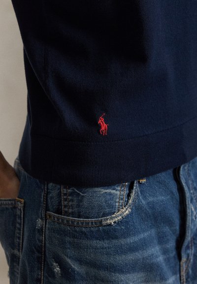 Gros plan d'une personne portant une chemise bleu marine avec un petit logo de cheval rouge, associée à un jean en denim bleu légèrement usé.