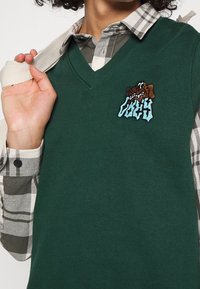 Grön bomullsärmlös tröja med v-ringning, med en broderad grafisk motiv, bärs över en rutig skjorta i grå och vita nyanser.