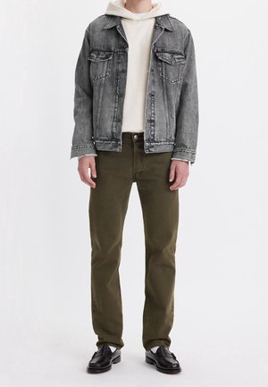 Denimjack in vervaagd zwart met een gestructureerd oppervlak, gedragen over een crèmekleurige hoodie. Olijfgroene broeken en zwarte instappers maken de look compleet.