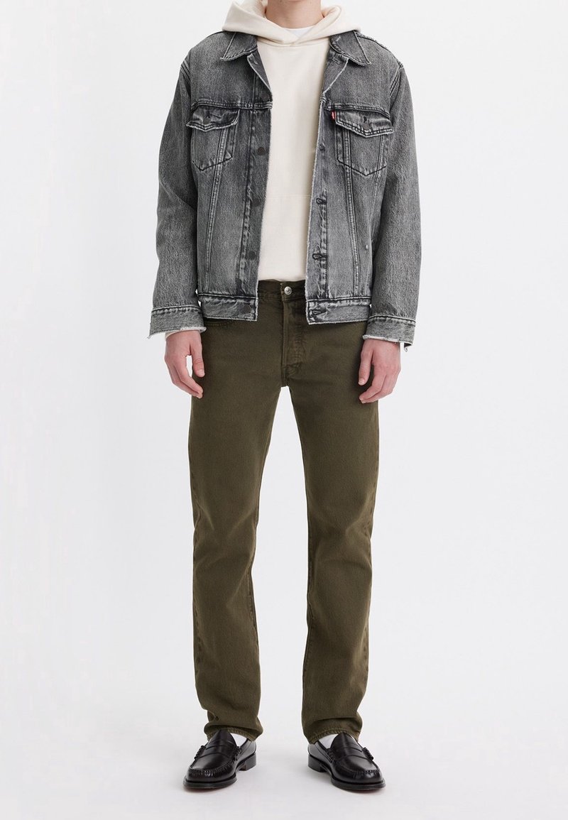 Džínová bunda ve světlé černé barvě s texturovaným povrchem, nošená přes krémovou mikinu. Zelené kalhoty a černé mokasíny dokončují vzhled.