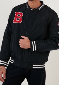 Schwarze Bomberjacke mit rotem "B"-Emblem, weiß gestreiftem, geripptem Kragen und Bündchen, Druckknöpfen, glatter Textur und elastischem Bund.