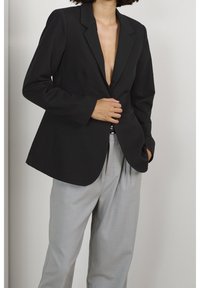Blazer nero su misura con rever a punta, chiusura a un bottone e silhouette slanciata, abbinato a pantaloni grigi chiari plissettati.