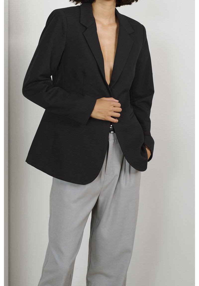 Blazer nero su misura con rever a punta, chiusura a un bottone e silhouette slanciata, abbinato a pantaloni grigi chiari plissettati.