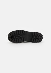 Semelle de chaussure en caoutchouc noir avec des crampons profonds et un logo embossé au centre, isolée sur un fond blanc uni.