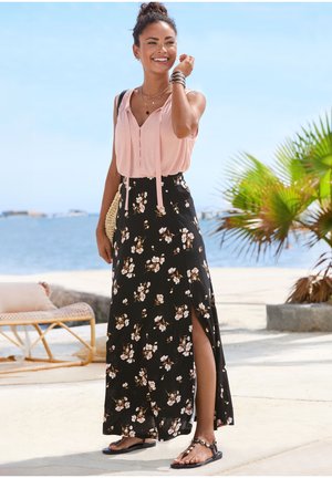 Smiling woman in pink sleeveless top and black floral maxi skirt with side slit stands on sunny beach near palm trees and wicker furniture.