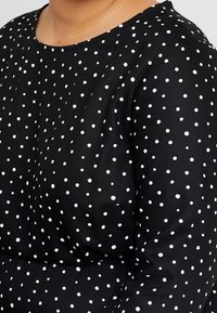 Schwarze Stoffbluse mit weißen Punkten, rundem Ausschnitt und glatter, strukturierten Oberfläche. Sie hat einen lässigen Schnitt und dezente Nähte.