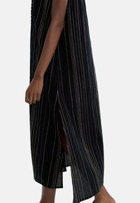 Czarna sukienka w pionowe, wielokolorowe paski, z bocznym rozcięciem i swobodnym krojem. Materiał wydaje się lekki i teksturowany.