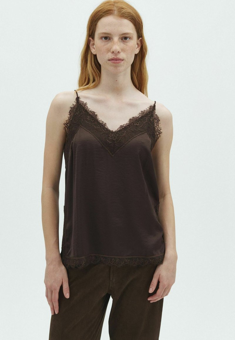 Femme rousse avec des taches de rousseur portant un caraco marron foncé bordé de dentelle et un pantalon foncé, debout devant un fond clair uni.
