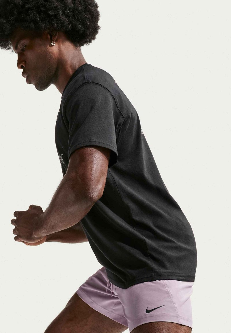 Homme avec coiffure afro et boucle d’oreille portant un t-shirt noir et un short Nike rose, penché en avant avec les mains jointes sur fond blanc.