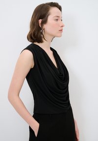 Schwarzes ärmelloses Oberteil mit drapierter Cowl-Nackenlinie, aus glattem Stoff und enger Silhouette. Getragen mit schwarzen Hosen, die Hände in den Taschen.