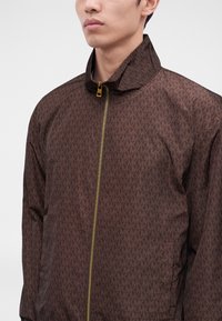 Homme portant une veste marron zippée avec un petit motif monogramme répété et un col relevé.