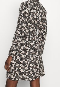 Květinové šaty s černým pozadím a bílými a oranžovými květinovými vzory. Dlouhé rukávy, nabíraný pas a krátký lem.