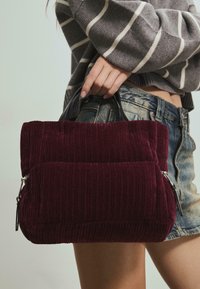 Burgunderfarvet corduroy håndtaske med to lynlåslommer, toppet med korte sorte håndtag. Tekstureret overflade med lodret ribbing.