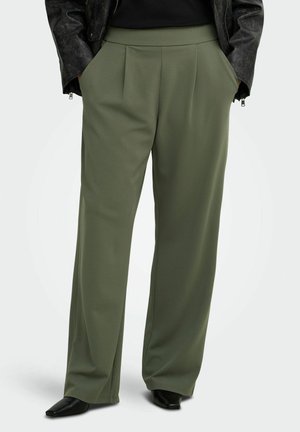 Personne portant un pantalon large vert olive, des chaussures noires pointues et une veste noire, les mains dans les poches.