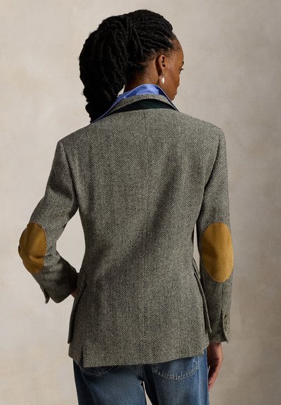 Femme aux cheveux tressés portant un blazer gris en chevrons avec des coudières beige et un jean bleu, vue de dos.