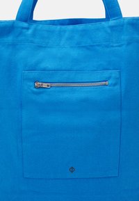 Bolsa de tela azul con un bolsillo frontal con cremallera, asas reforzadas y un logotipo negro en la esquina inferior derecha.