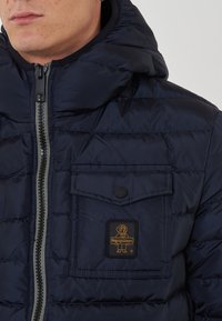 Giubbotto imbottito blu marina con chiusura a zip, design trapuntato e tessuto testurizzato. Presenta una tasca frontale con bottone a scatto e etichetta del marchio.