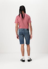 Rdeča majica s kratkimi rokavi v kombinaciji z modrimi denim kratkimi hlačami z žepi, ravnim krojem in obrabljenim videzom. Oblečeni so črno-beli športni copati.