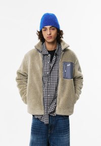 Mlad moški z kodrastimi lasmi, oblečen v modro pleteno kapo, bež polarno jakno, srajco v črno-beli karirasti vzorec in modre kavbojke, roke v žepih.