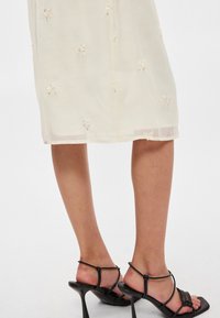 Vestido creme com bordados florais, material transparente, até ao joelho. Combinado com sandálias de salto alto pretas com tiras. Textura suave.