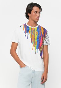 Mann iført hvit T-skjorte med fargerik, dryppende geometrisk mønster og trykt tekst på høyre erme, kombinert med lyseblå jeans.