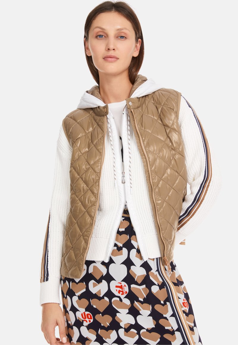 Quilted beige puffer vest with a hood worn over a white knit sweater. The ensemble features a patterned skirt with hearts in brown and white.