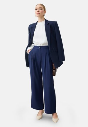 Kvinde iført en navy blazer kastet over skuldrene, hvid bluse, navy vide bukser, beige hæle og holder en lille brun håndtaske.