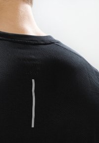 Camiseta deportiva negra con una tela texturizada, que presenta un detalle de franja reflectante vertical en la parte superior de la espalda y un cuello redondeado.