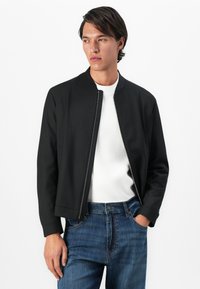 Joven vestido con chaqueta negra con cremallera sobre una camisa blanca y jeans azules, de pie con una mano en el bolsillo, contra un fondo blanco.