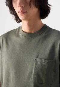 T-shirt en coton vert olive avec un col rond, des manches courtes et une poche poitrine avec des détails de coutures décoratives.