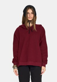 Młoda kobieta w ciemnoczerwonej, obszernej bluzie z kapturem, czarnej czapce związanej pod brodą, czarnych spodniach oraz z wieloma srebrnymi bransoletkami na lewej ręce.