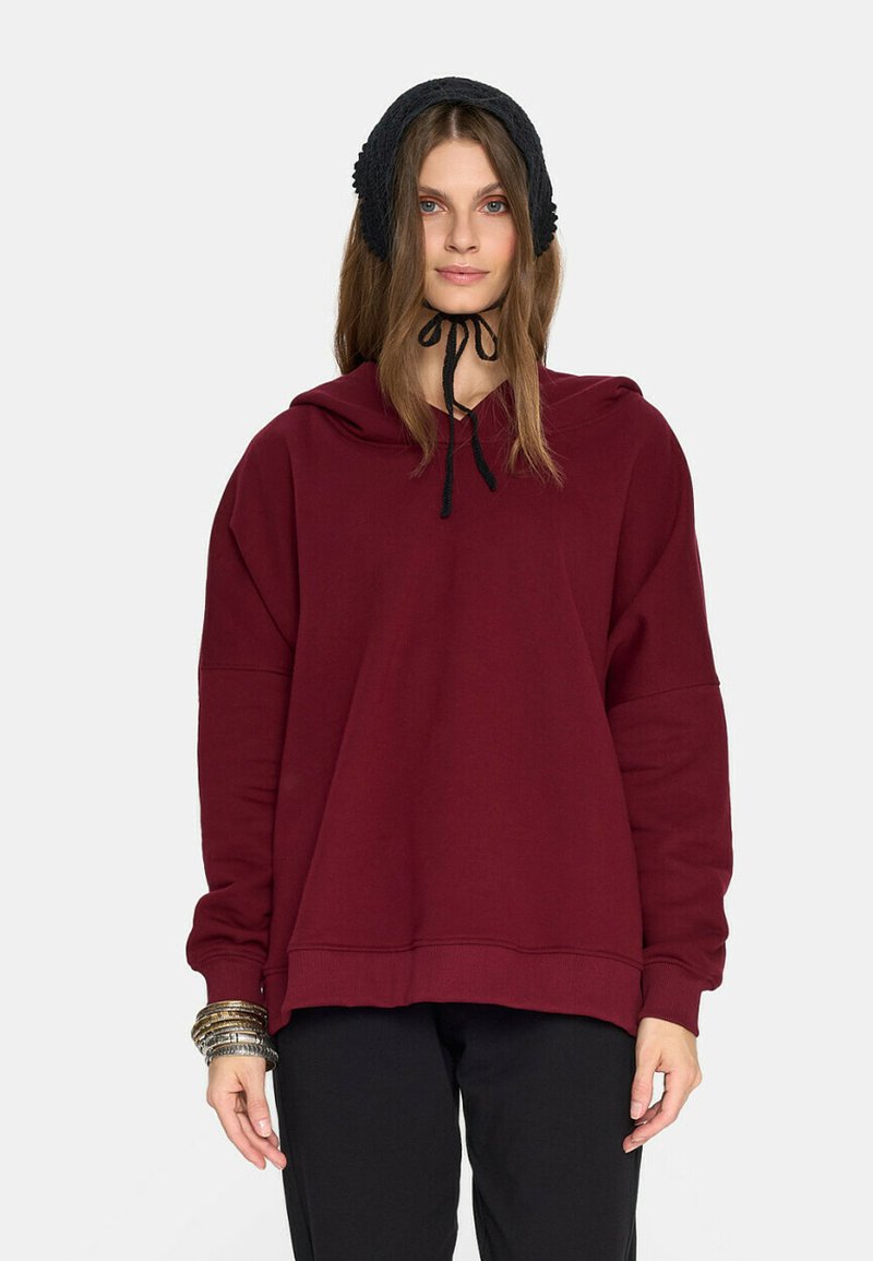 Młoda kobieta w ciemnoczerwonej, obszernej bluzie z kapturem, czarnej czapce związanej pod brodą, czarnych spodniach oraz z wieloma srebrnymi bransoletkami na lewej ręce.