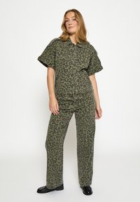 Olívovo zelený leopardí vzor dvojdielneho oblečenia s krátkym rukávom, zapínaným na gombíky, a širokými nohavicami vyrobenými z mäkkej látky. Čierne topánky.