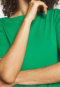 Grön kortärmad t-shirt i mjukt tyg med båtkrage. Bärarens armar är korsade, vilket visar den naturliga hudens textur.
