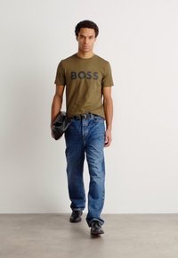 Alyvuogių žalios medvilnės marškinėliai su "BOSS" logotipu, derinami su mėlynais džinsais ir juodais batais. Modelis laiko juodą krepšį, stovėdamas ant pilko grindų.