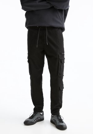 Pantalon cargo noir en coton avec plusieurs poches latérales, taille à cordon de serrage et coupe slim, associé à des baskets grises.