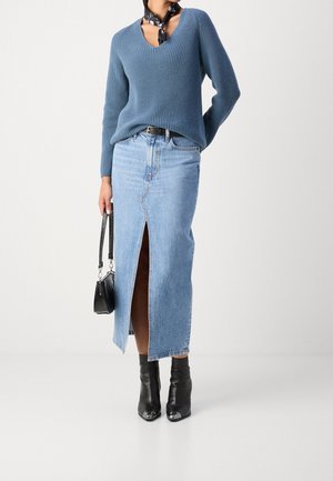 Femme portant un pull bleu col V, une jupe en jean bleu clair avec une fente à l’avant, des bottines noires, une ceinture noire, une écharpe, et tenant un sac à main noir.