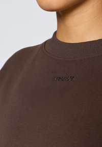 Close-up de um suéter castanho de gola redonda com colarinho canelado e a palavra "STRONGER" bordada no peito, usado por uma pessoa.