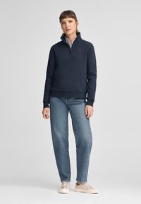 Pullover navy in misto cotone, con collo a zip a un quarto e accento del logo rosso. Abbinato a jeans azzurri a gamba dritta e sneakers beige.