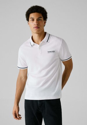 Camisa polo blanca con ribete negro en el cuello y las mangas, con un pequeño logo negro de Red Bull Racing en el lado izquierdo del pecho, combinada con jeans oscuros.