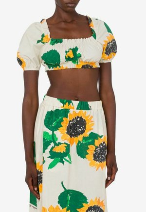 Donna che indossa un completo due pezzi con maniche corte a sbuffo e stampa di girasoli su tessuto bianco, mostrando l'addome e la vita della gonna.
