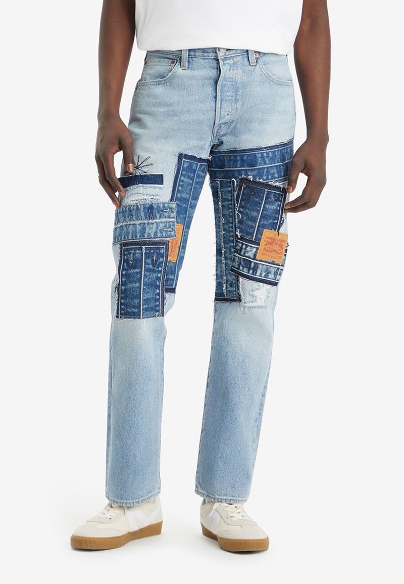 Personne portant un jean bleu clair avec des patchs en denim bleu foncé et des bords effilochés, associé à des baskets blanches et un haut blanc.