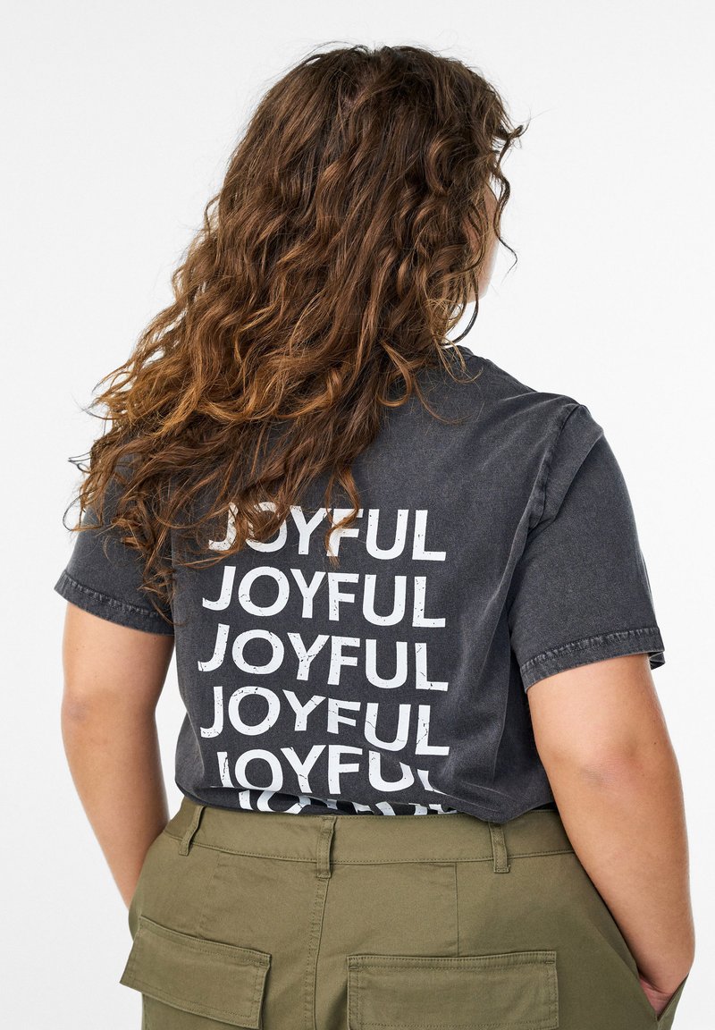 Persona ar cirtainiem brūniem matiem, kas valkā izbalējušu melnu T-kreklu ar uzrakstu "JOYFUL" baltā krāsā atkārtoti uzdrukātu uz muguras un olīvu zaļās bikses.