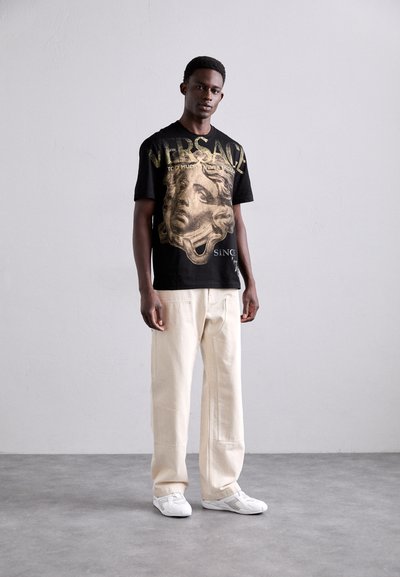 T-shirt en coton noir avec impression graphique dorée d'un visage et de texte ; associé à un pantalon large beige et des baskets blanches.