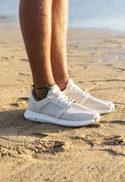 Baskets de sport beige clair avec des panneaux en maille, semelles et lacets blancs, reposant sur un sol sablonneux près de l'eau, mettant en avant un design simple.