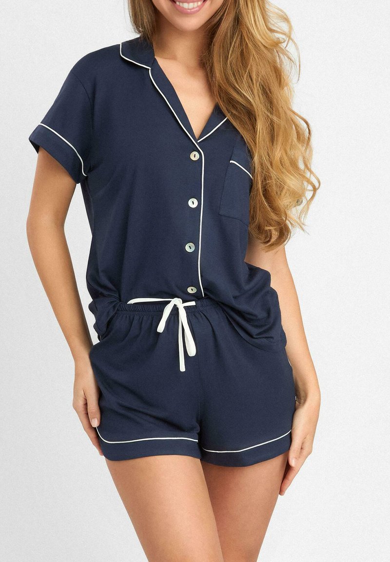 Granatowy komplet piżamowy z miękkiego materiału, składający się z bluzki z krótkim rękawem z białymi lamówkami, guzikami i kieszonką, w komplecie z szortami na elastycznej talii.