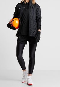 Sort atletikjakke med lynlås, sort skjorte, sorte shorts, sorte leggings, handsker og hvide sko; holder en orange fodbold.