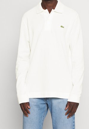 Mand iført en hvid langærmet poloshirt med et lille grønt logo på brystet, kombineret med lyseblå jeans.