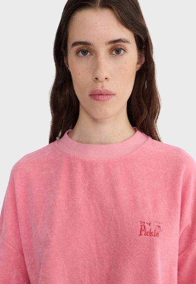 Jeune femme aux longs cheveux bruns portant un sweat-shirt rose à col rond avec un petit logo "The Dog Pickle" sur la poitrine.