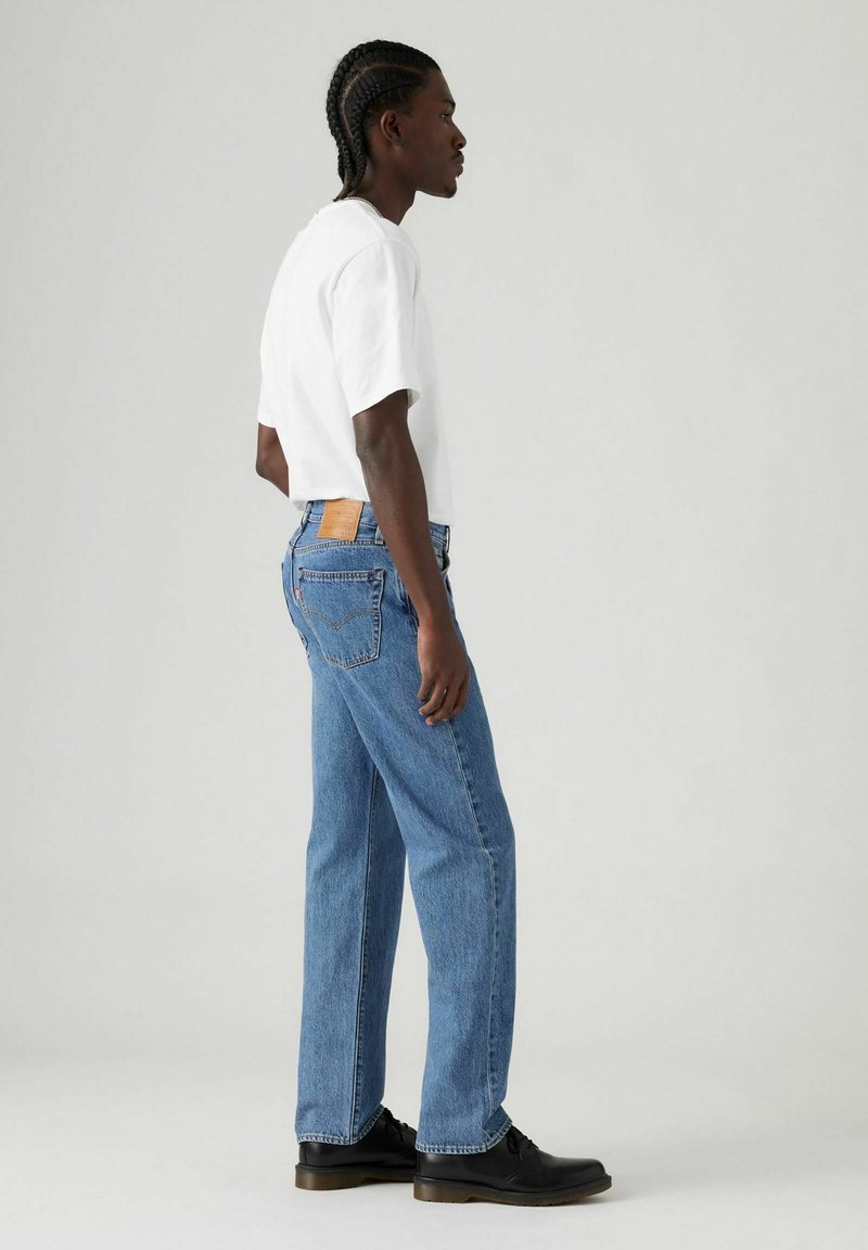 Vaqueros azules de mezclilla con un corte recto, con un parche de cuero marrón en la parte trasera, combinados con una camiseta blanca de manga corta y zapatos negros.